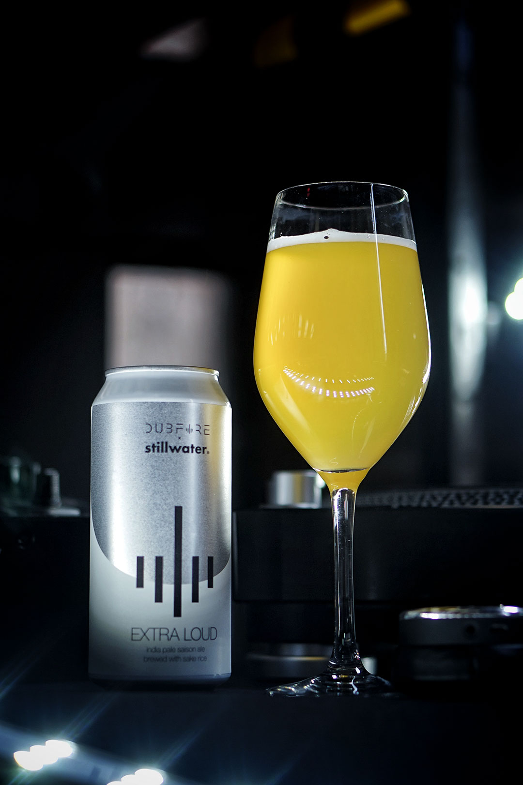 An image of Extra Loud in a stemmed beer glass with the can and dj equipment on a dark background